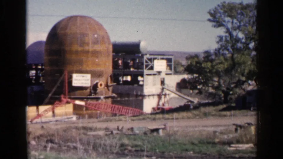 1960 Sioux Falls-area 8mm frame with a rusted dome-like structure, nearby scaffolding, fencing, signage, and a tree at the edge of the site.