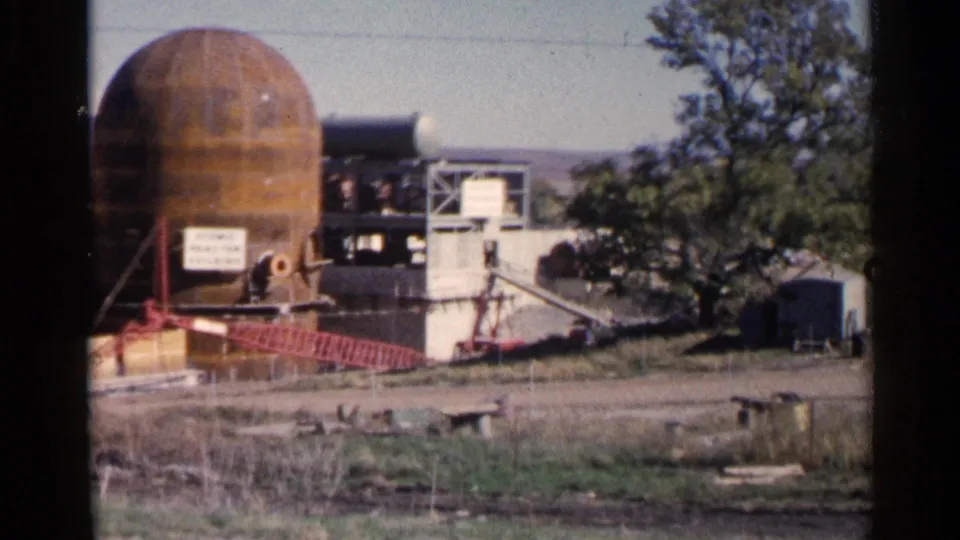 1960 Sioux Falls-area home-movie frame panning across the rusted industrial dome, adjacent work platform, fencing, and roadside ground.