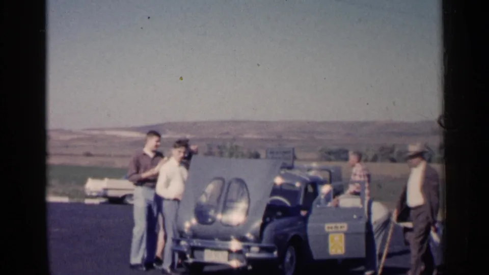 1960 South Dakota home-movie frame showing several men near a blue car with its hood raised against open fields and distant hills.