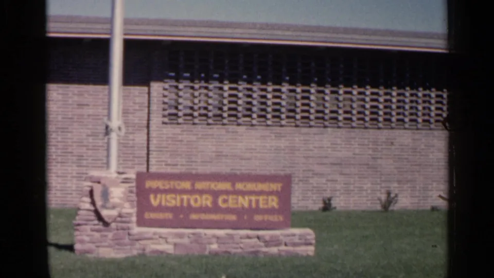 The Pipestone National Monument Visitor Center sign remains framed with a flagpole, stone base, grass and mid-century brick facade.