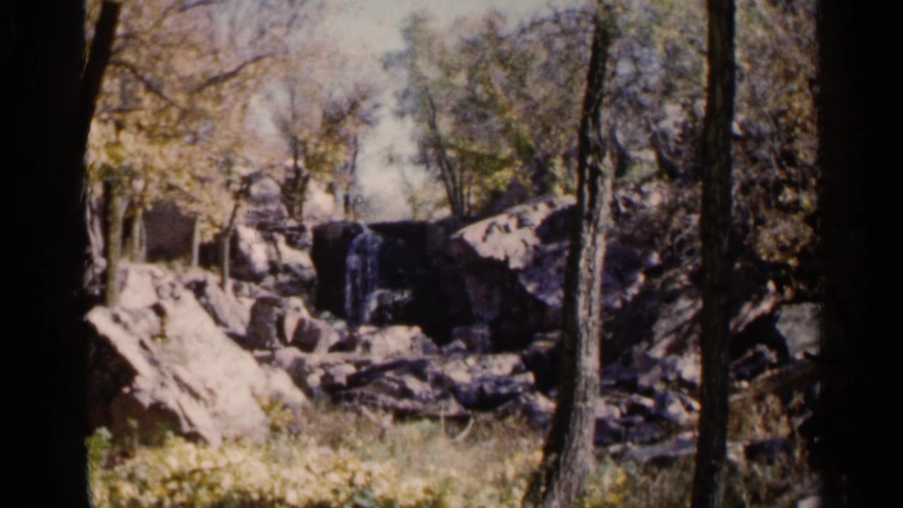 Small waterfall over rocks seen through trees in a wooded park setting.
