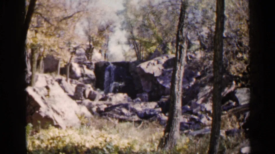 1960 8mm frame showing a small South Dakota waterfall, pale rocks, tree trunks, and patchy autumn vegetation.