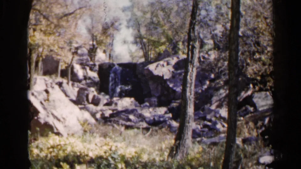 1960 8mm South Dakota park scene with a small waterfall, rocky ledge, trees, and a seated adult partly visible near the right edge.