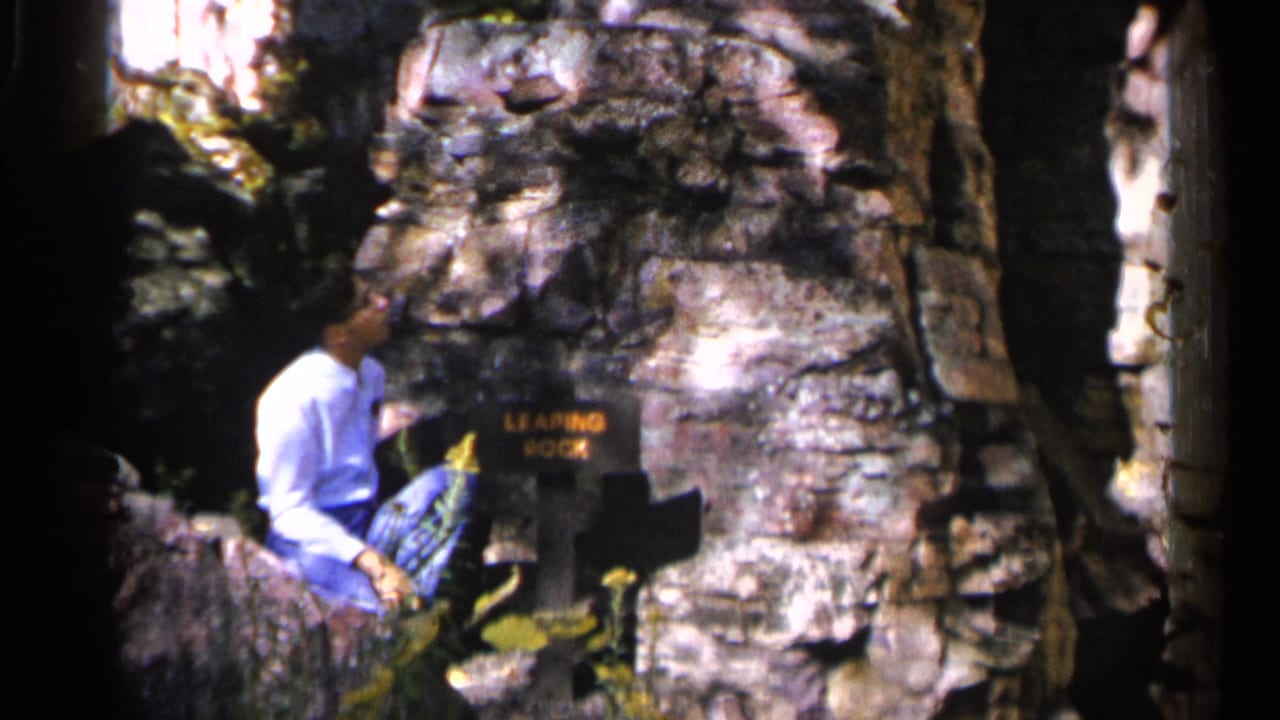 The visitor remains seated by the Leaping Rock sign, with layered boulders and tree shade visible around him in Sioux Falls.