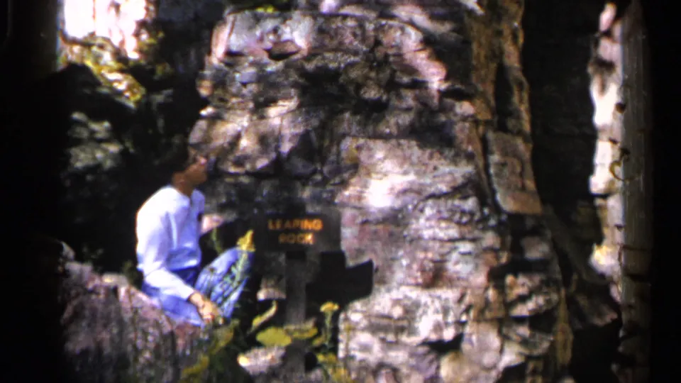 The visitor remains seated by the Leaping Rock sign, with layered boulders and tree shade visible around him in Sioux Falls.