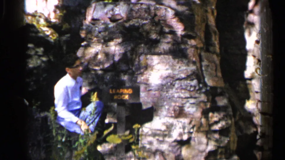 A close home-movie view shows the man resting beside the Leaping Rock marker amid boulders, leaves, and mottled sunlight.