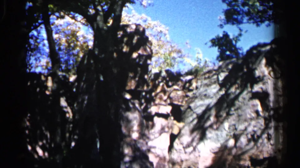 The scene shifts to a wider view of the rocky landscape, dark tree silhouettes, foliage, and bright blue sky in Sioux Falls.