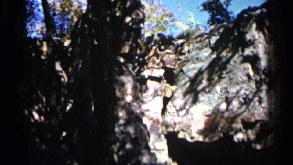 A 1960 outdoor 8mm frame shows a rugged grey rock face viewed through trees and high-contrast shade.
