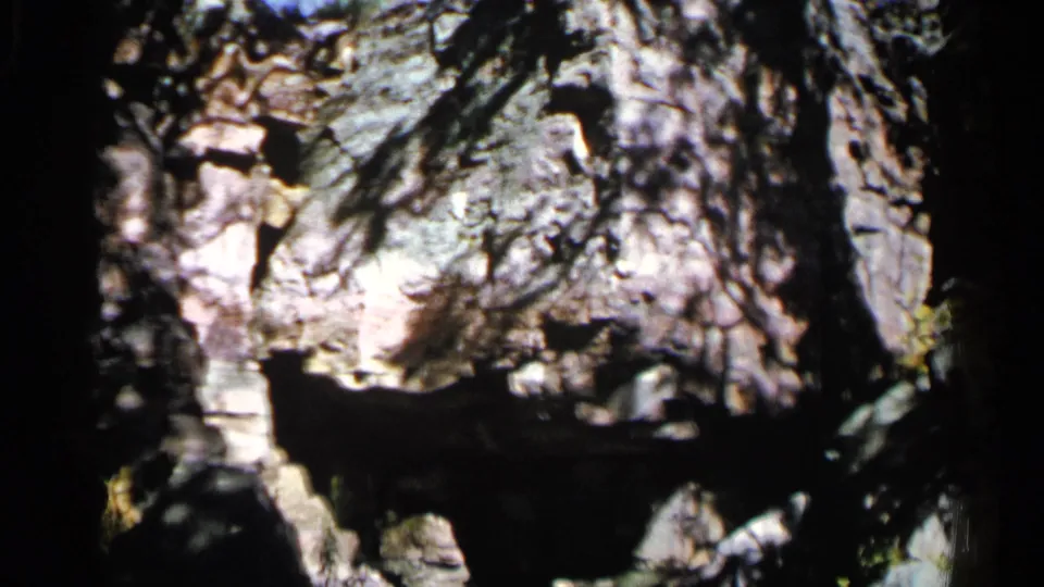 1960 8mm frame from Sioux Falls showing a wooded rock face, heavy shadows, and bright sunlight across the stone.