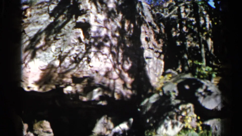 1960 8mm frame from Sioux Falls with a shadowed cave edge, textured rock, tree shadows, and greenery near the opening.