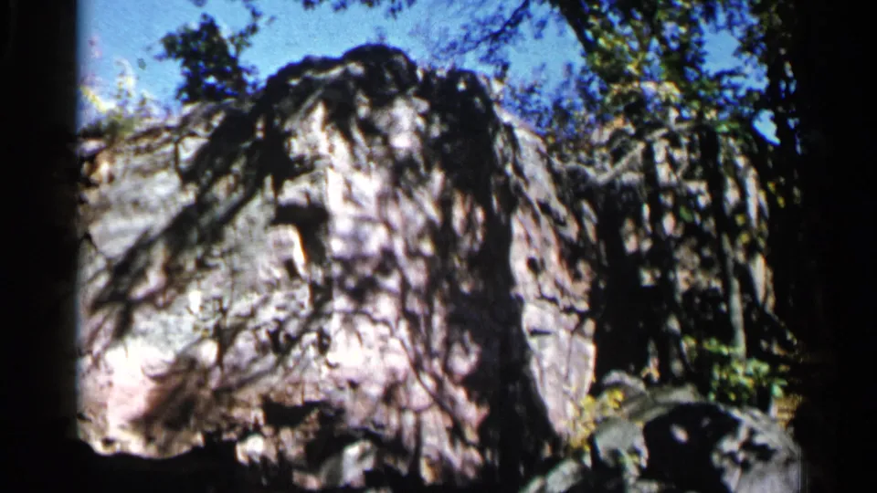 1960 8mm frame from Sioux Falls looking upward from a dark opening toward a rock face, blue sky, and trees.