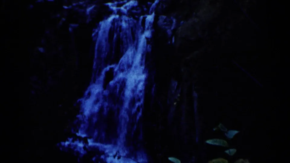 1960 8mm frame from Sioux Falls showing a waterfall tinted deep blue against a dark rock face, with leaves at the lower edge.