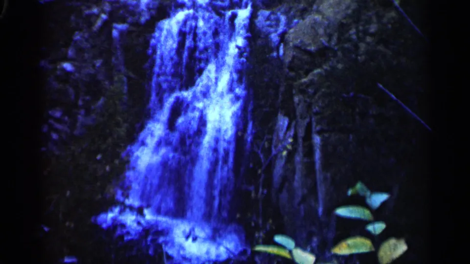 1960 8mm frame from Sioux Falls showing a blue-tinted waterfall descending over dark rocks with green leaves in the foreground.