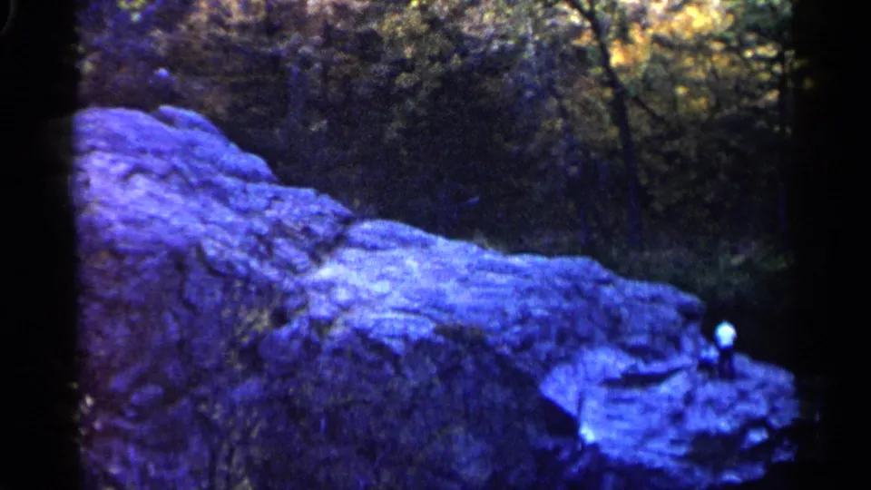 1960 8mm view in Sioux Falls with a distant visitor on a blue-purple rocky ledge beneath dark trees.