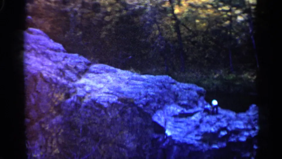 1960 8mm frame from Sioux Falls showing a lone hatted person on a rocky outcrop in a wooded waterfall setting.