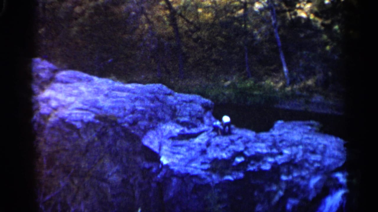 Person standing on a rock outcrop above a small dark pond with trees behind.