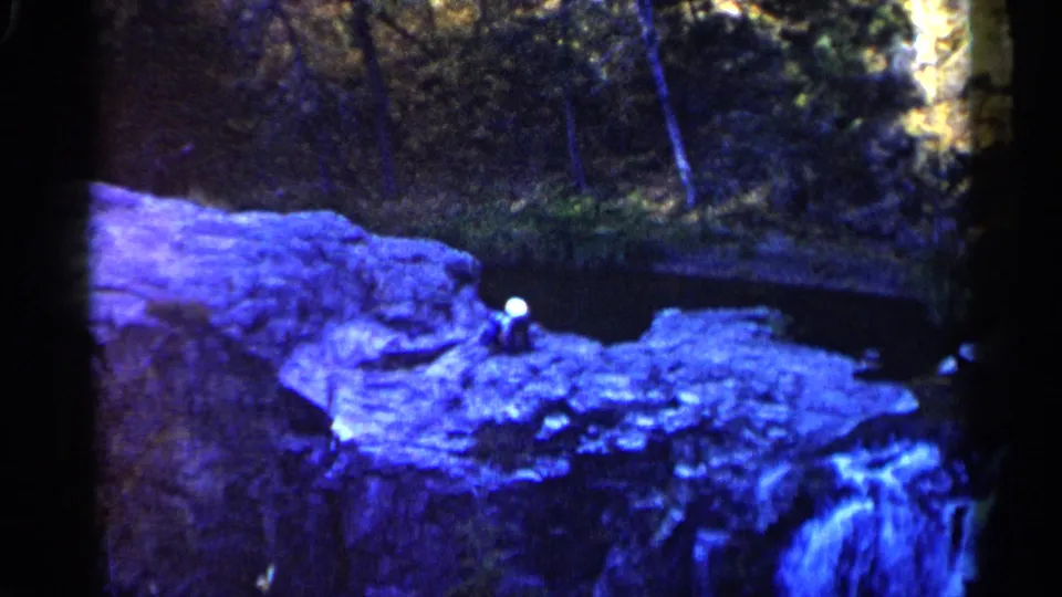 1960 Sioux Falls home-movie frame showing a person on a rock ledge above dark water with a small waterfall visible below.