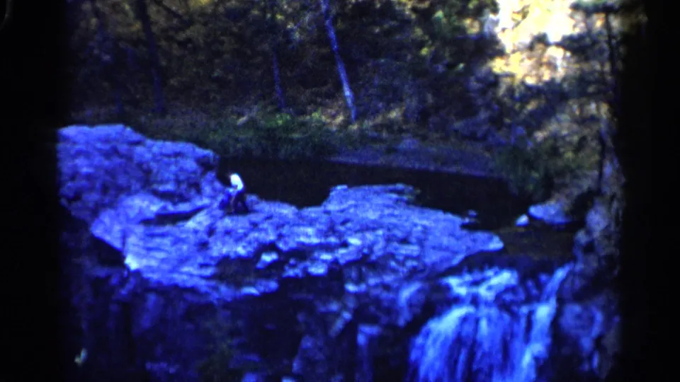 1960 Sioux Falls frame showing a person on a rock formation with pond water behind and a small waterfall at the lower right.