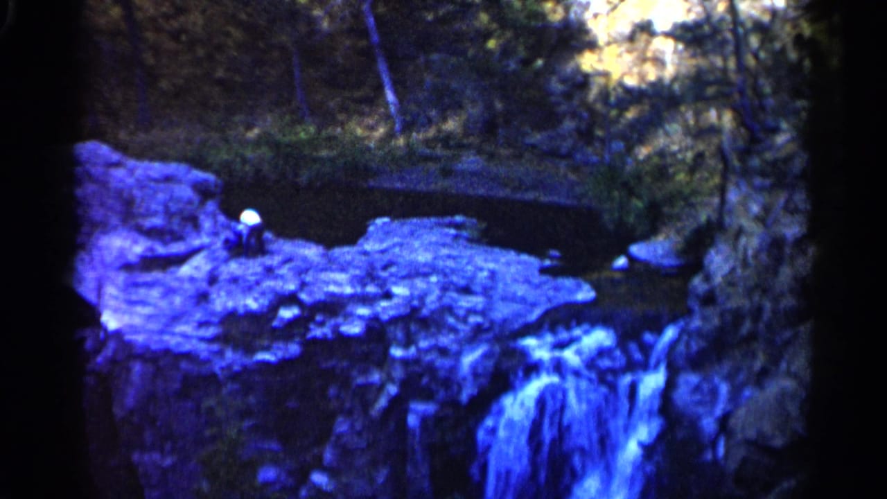 Blue-tinted waterfall over rocks in a wooded area with a small seated figure near the upper ledge.