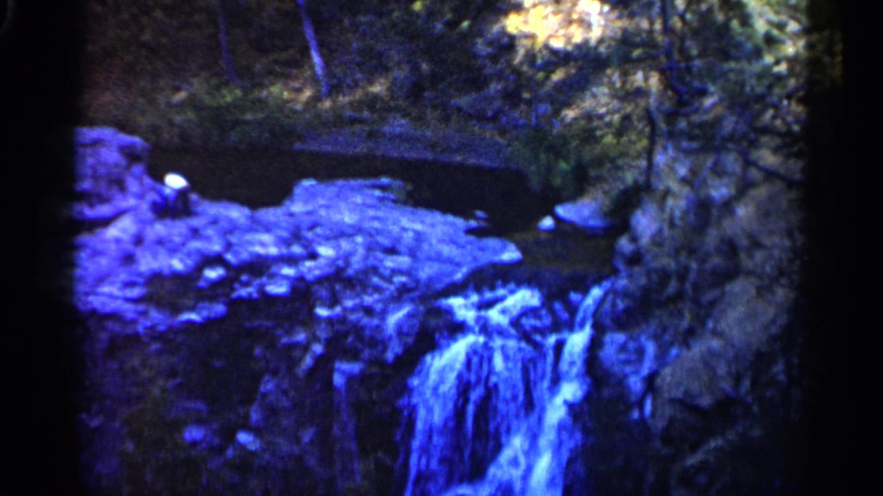 Blue-cast 1960 Sioux Falls waterfall frame with rocky ledges, wooded shade, falling water, and a small seated visitor near the top.