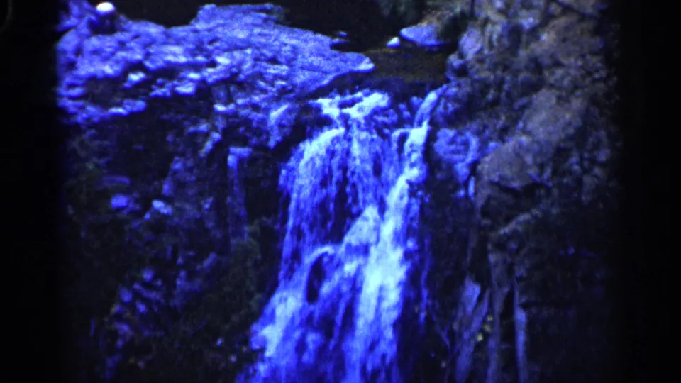 Final 1960 Sioux Falls 8mm frame showing a shaded rocky waterfall, wooded banks, strong blue color shift, and a small seated figure near the top.