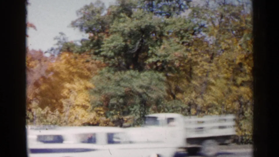 A white mid-century car passes a tree-lined road in the 1960 Sioux Falls autumn home-movie footage.