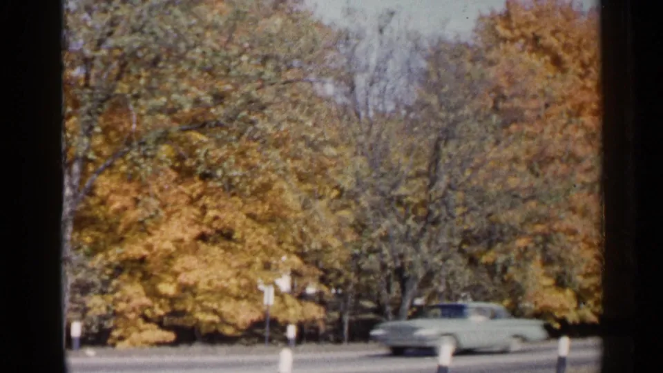 1960 Sioux Falls frame showing a light-colored station wagon moving past fall trees on a paved roadside.