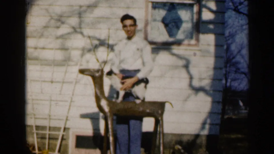 A young man in glasses and a white shirt holds a black-and-white cat near a wooden deer statue outside a white-sided Sioux Falls house in 1960.