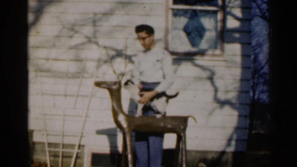 The young man stands by the white-sided house, holding the cat over or near the wooden deer statue in this 1960 Sioux Falls home-movie frame.