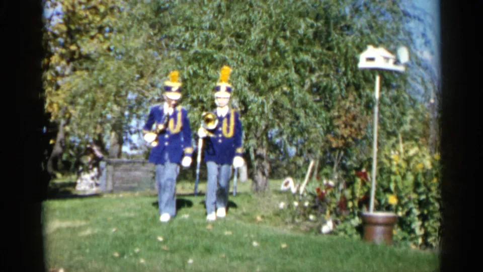Two young band members in blue uniforms march through a yard in the supplied 1960 Sioux Falls setting.
