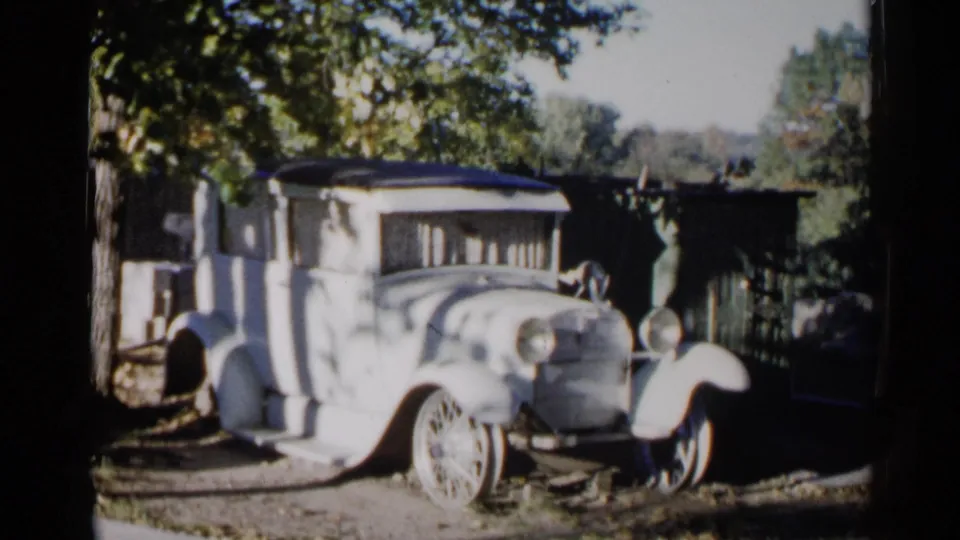 1960 Sioux Falls 8mm frame showing the side-front of a light antique car in dappled shade outdoors.