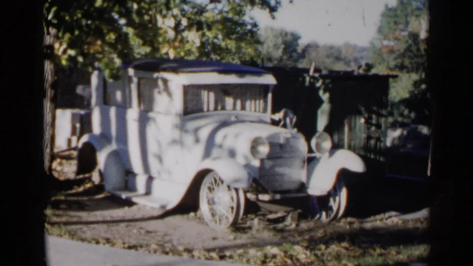 1960 Sioux Falls home-movie still of a light-colored antique car framed by trees and strong sun-shadow contrast.