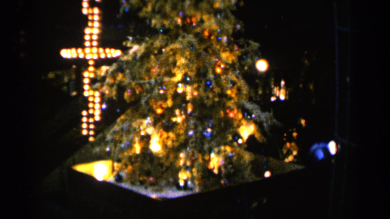 Decorated Christmas tree with warm lights beside a glowing bulb cross at night.