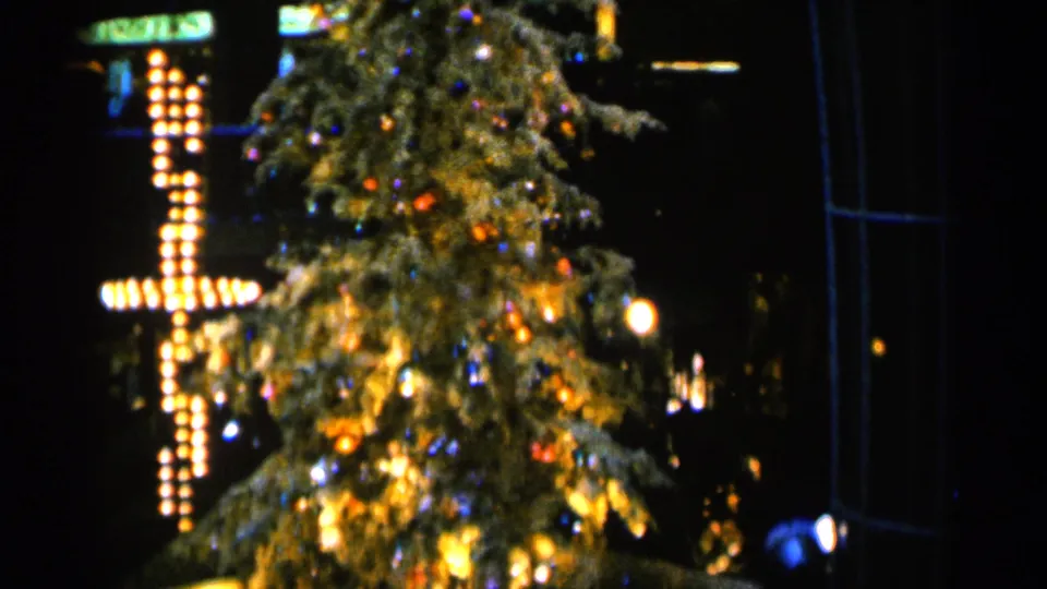 1960 Sioux Falls frame showing the lower view of a decorated Christmas tree with a bright bulb cross and dark surroundings.