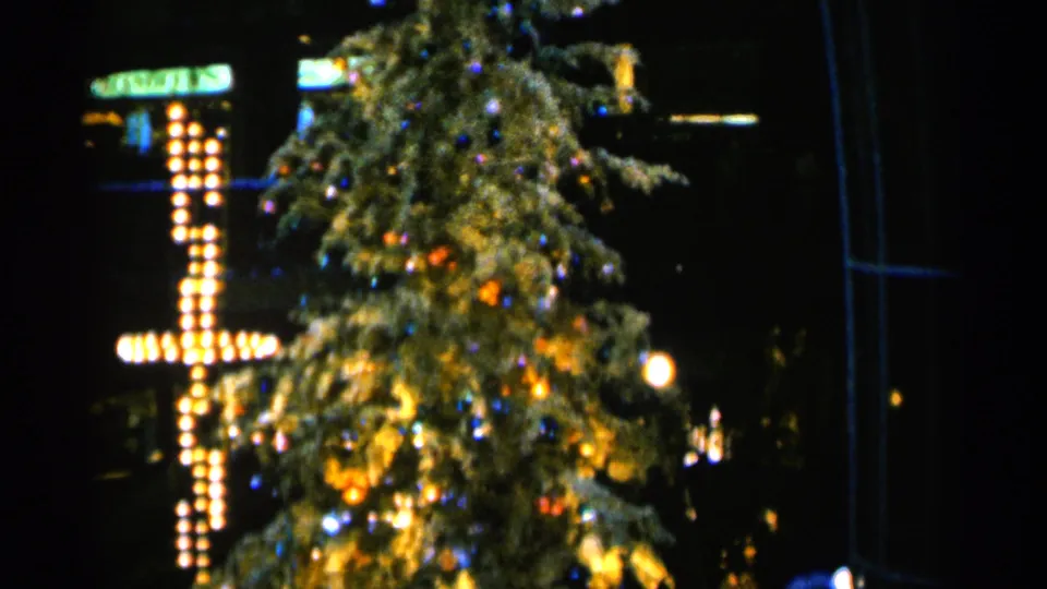 1960 Sioux Falls 8mm frame with a softly focused Christmas tree, colorful ornaments, and a glowing bulb cross at night.
