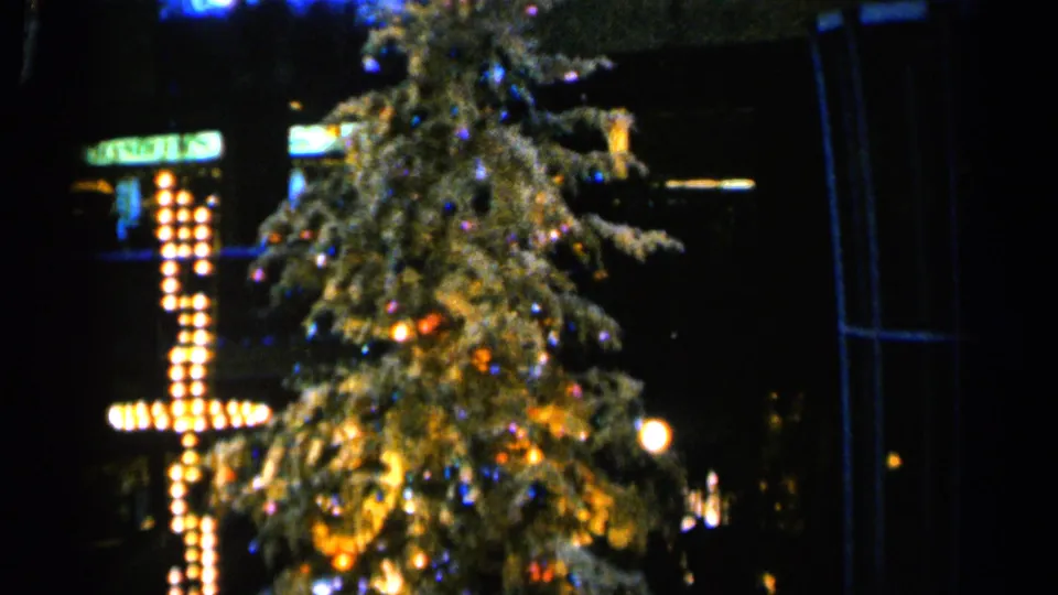 1960 Sioux Falls 8mm frame showing a lighted Christmas tree with ornaments and a bulb-lit cross in a dark outdoor setting.