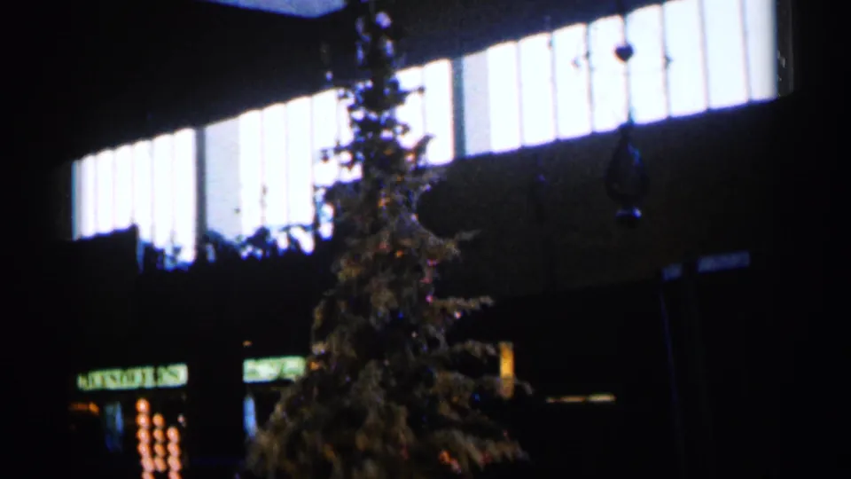 Soft 1960 Sioux Falls 8mm frame of a Christmas tree in a dim public-looking interior with a partial sign at lower left.