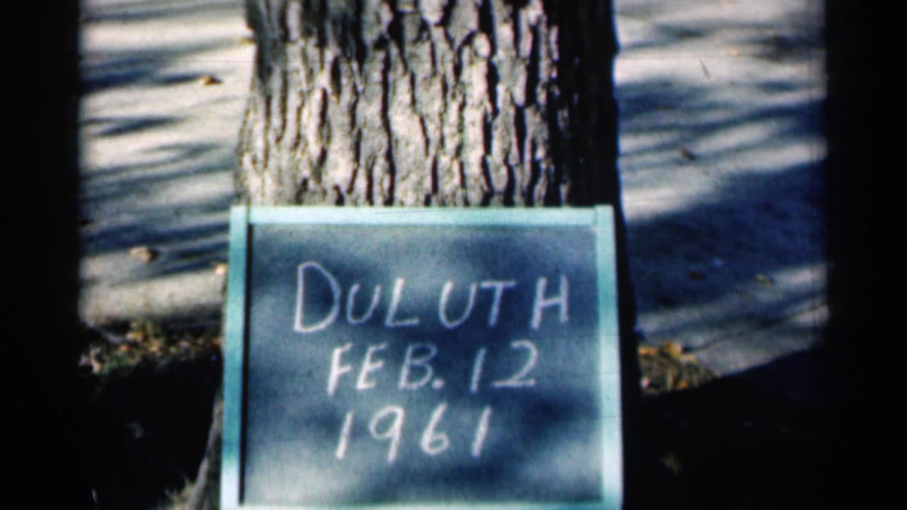 Duluth, Minnesota 1961 home-movie frame with a handwritten chalkboard date sign leaning against a tree in winter light.