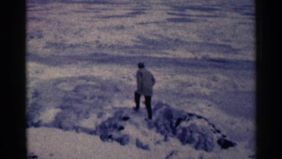 A bundled adult appears farther along the snowy edge in the 1961 Duluth home-movie scene, seen from a distance.