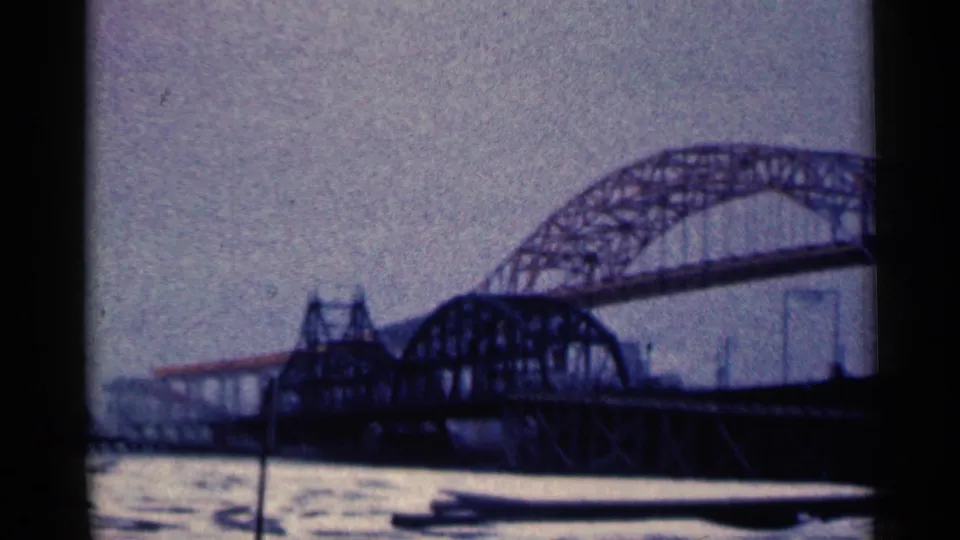 1961 Duluth frame showing the arched metal truss bridge across gray water, with distant shoreline features under a pale sky.