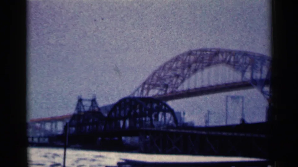 1961 Duluth home-movie frame showing a large steel arch truss bridge spanning river water under an overcast sky.