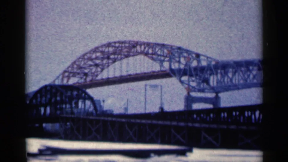 1961 Duluth home-movie frame showing a steel arch bridge behind a darker foreground truss bridge over water.