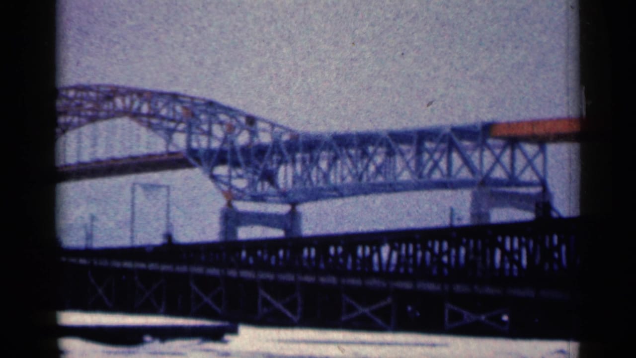 Two metal bridges overlap above water, with a dark truss bridge in front and a pale arched bridge behind.