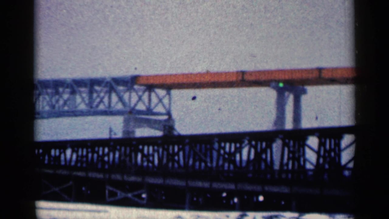 Snowy construction scene with dark bridgework, orange elevated beam, and concrete supports.