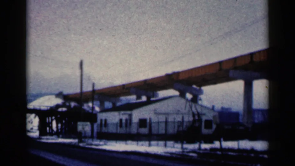 1961 Duluth home-movie frame of a fenced white building, utility poles, snow, and a sloping elevated beam overhead.