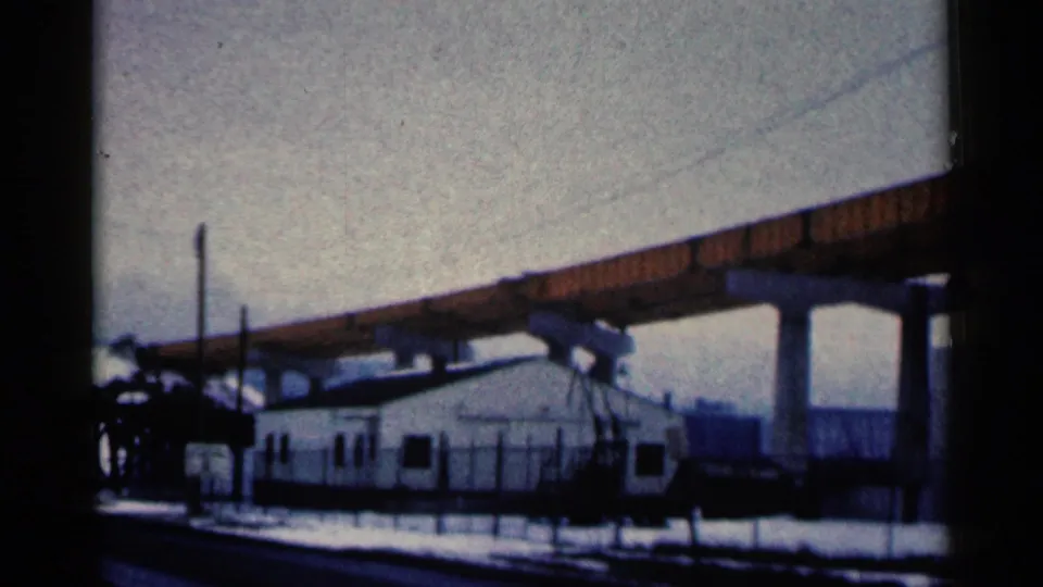 1961 Duluth 8mm frame showing a white building behind fencing, snowy ground, concrete supports, and elevated bridge beams.