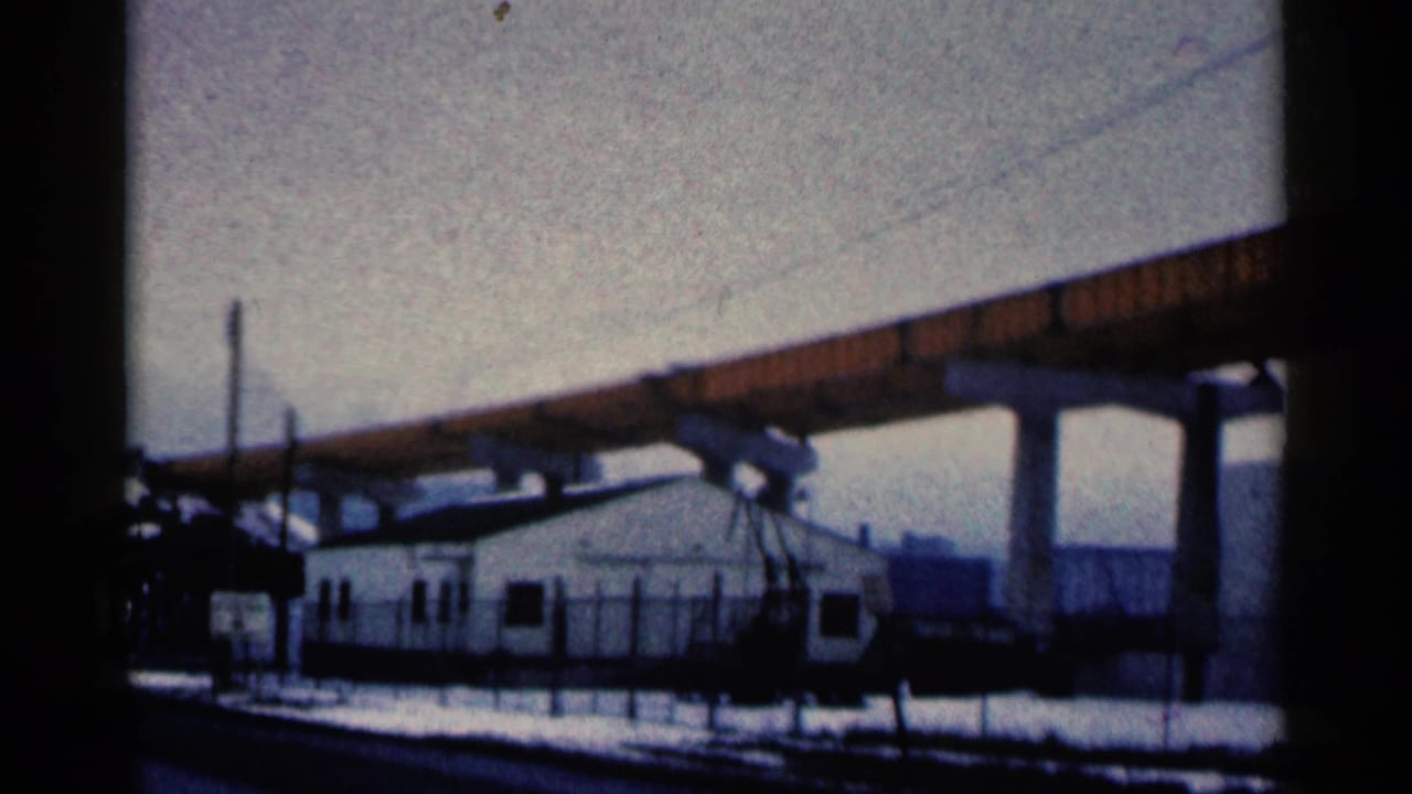 1961 Duluth home-movie frame showing an orange elevated bridge over a small white building with snowy ground and tanks beyond.