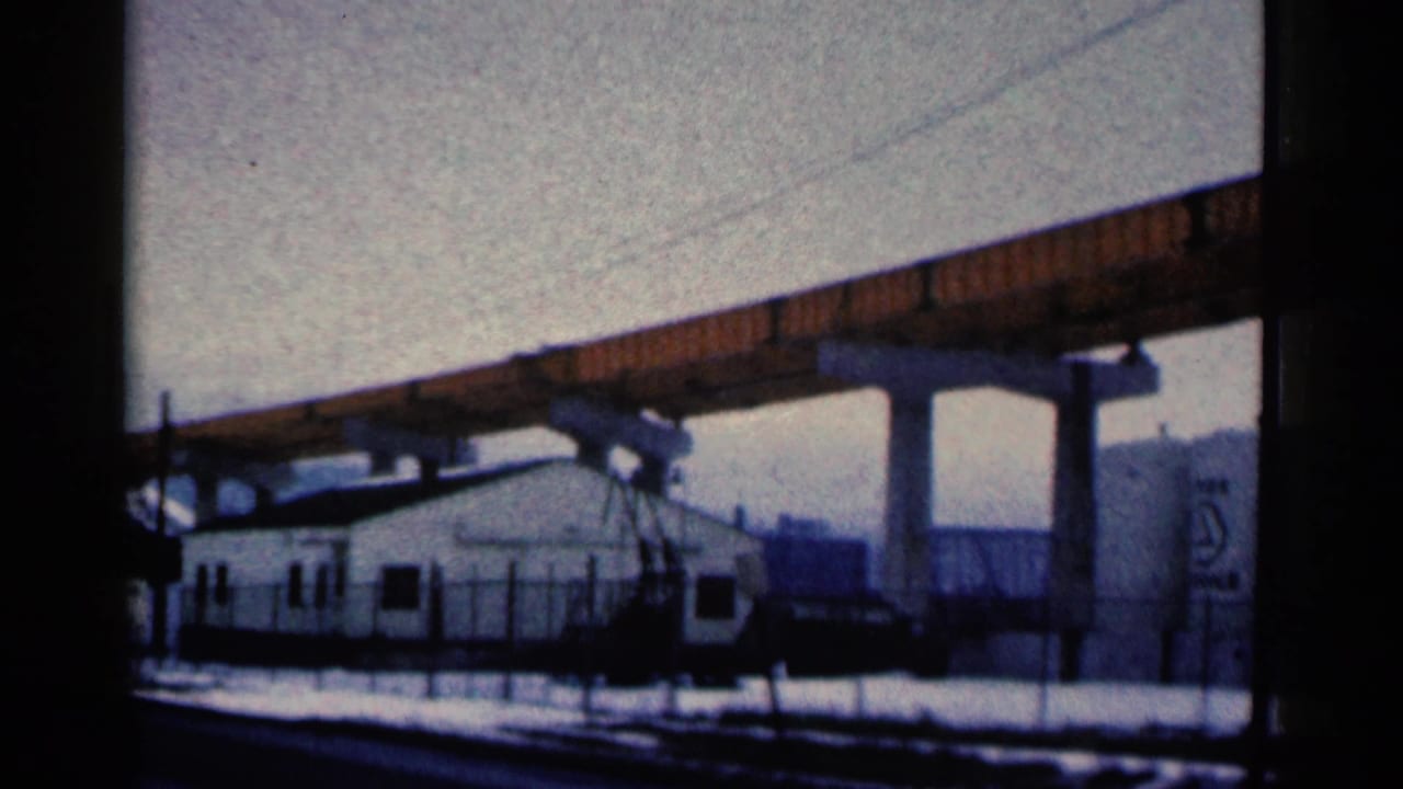 1961 Duluth frame with a large orange bridge crossing over a white building, snowy fenced ground, and a gray tank with a marking.