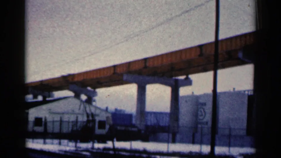 1961 Duluth home-movie frame showing bridge supports above a small white building, snowy fenced ground, and large industrial tanks.