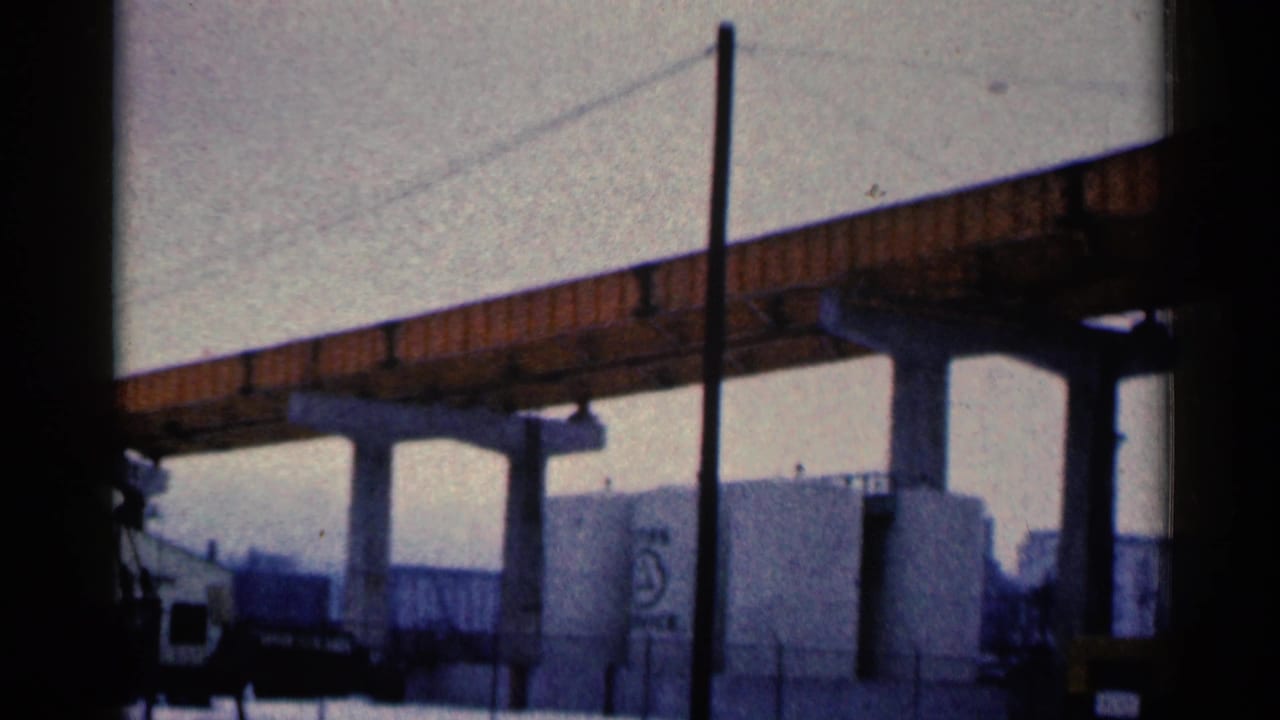 Orange steel bridge over concrete supports and storage tanks in an industrial area.